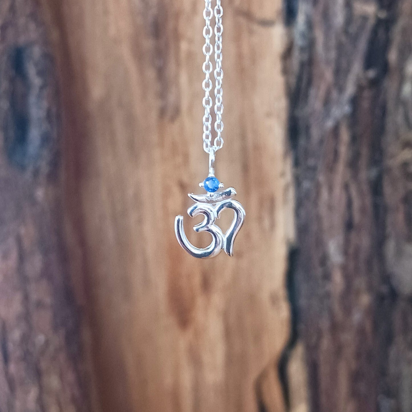 This lovely Om Symbol silver pendant is very dainty and comes with an inlaid Corundum blue stone, on a fine solid 925 silver chain. Om or (Aum) is a sacred sound considered by many ancient philosophical texts to be the sound of the universe, encompassing all other sounds within it. In Sanskrit, om is called Pranava, which means to hum, and is considered an unlimited or eternal sound.