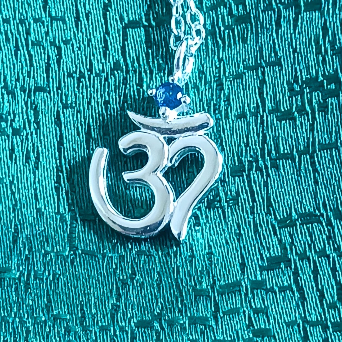 This lovely Om Symbol silver pendant is very dainty and comes with an inlaid Corundum blue stone, on a fine solid 925 silver chain. Om or (Aum) is a sacred sound considered by many ancient philosophical texts to be the sound of the universe, encompassing all other sounds within it. In Sanskrit, om is called Pranava, which means to hum, and is considered an unlimited or eternal sound.
