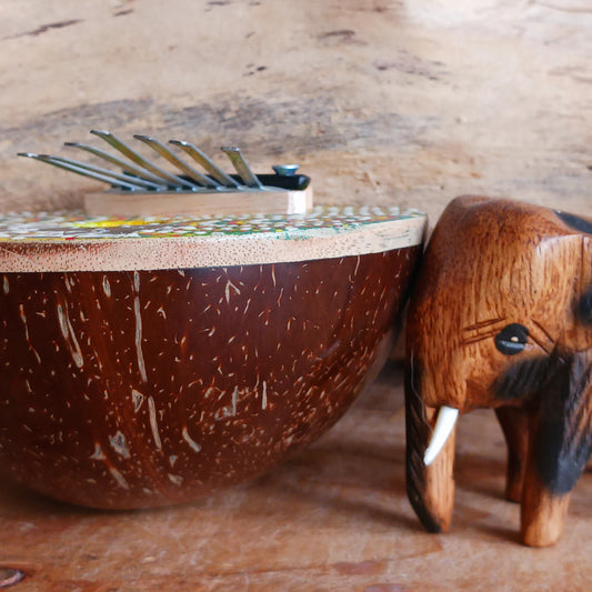 This thumb piano has been hand carved out of coconut shell and decorated with vibrant colour dot painting of an elephant. The piano board itself has been placed over a polished half coconut, which creates a resonating chamber that produces a louder, more harmonically complex sound. It has 7 keys which are adjustable and easy to tune by tightening or loosening the screws on the top.