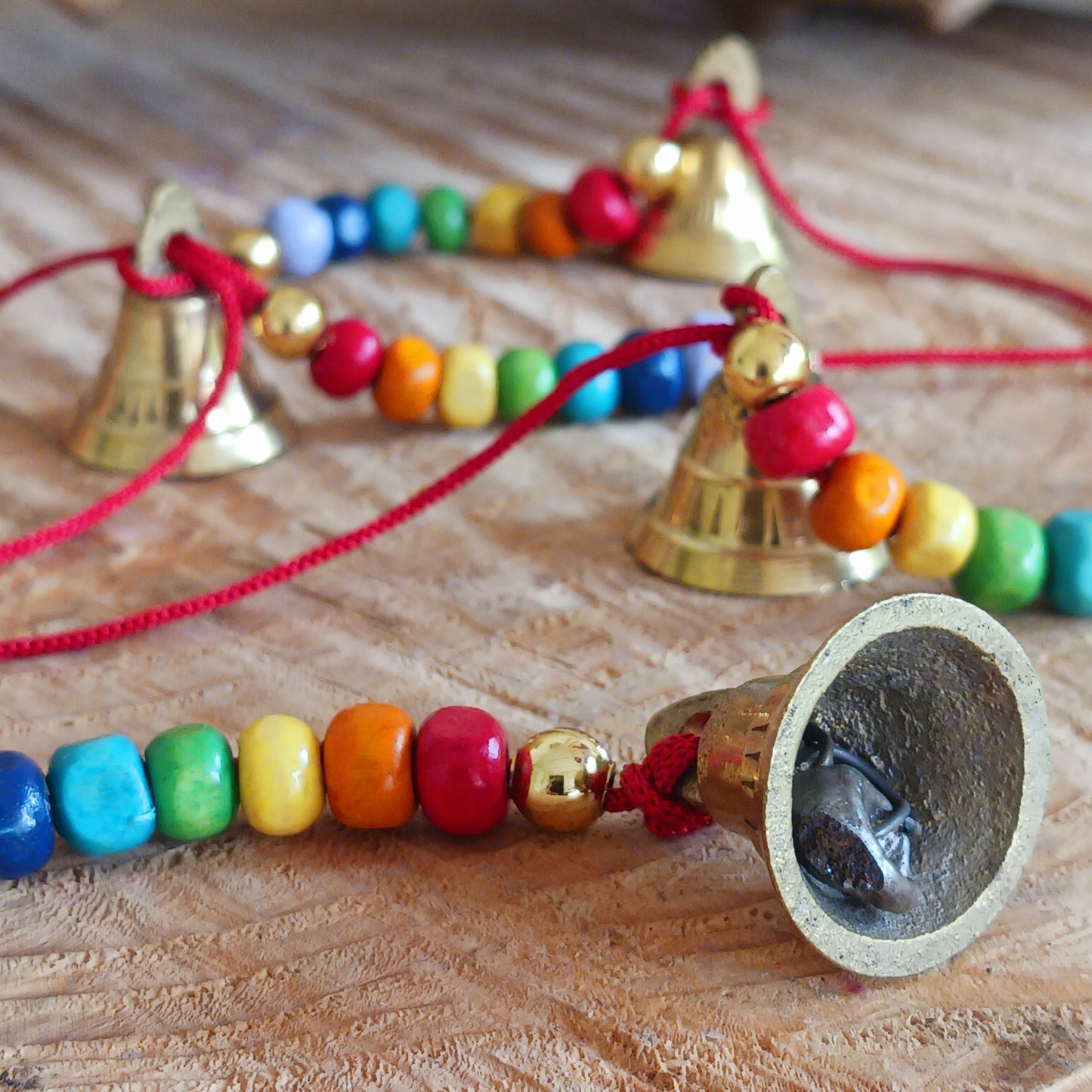 This delightful string of five Indian brass bells, hung on a length of cord have vibrant rainbow colour wooden beads. This lovely little string of bells makes a wonderful gentle jingle sound when hung in the breeze.