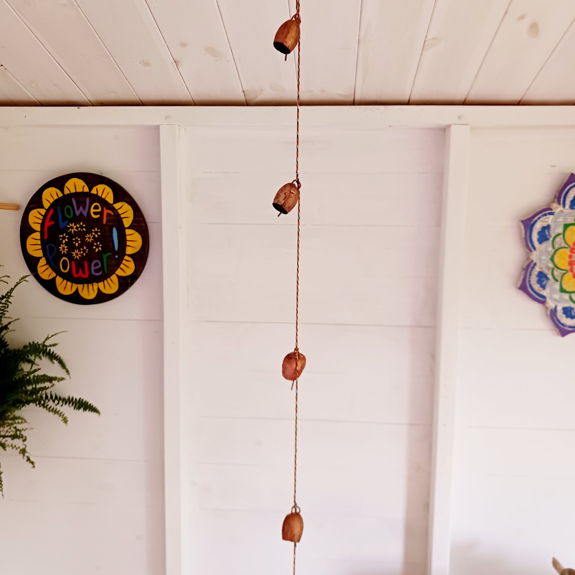 String of small rustic handmade ironwork bells with copper coloured paintwork, hung on a length of colourful string. Each bell has a small metal clapper which produces a gentle tinkling sound.