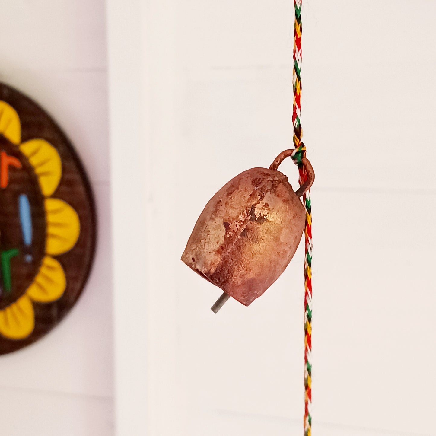 String of small rustic handmade ironwork bells with copper coloured paintwork, hung on a length of colourful string. Each bell has a small metal clapper which produces a gentle tinkling sound.