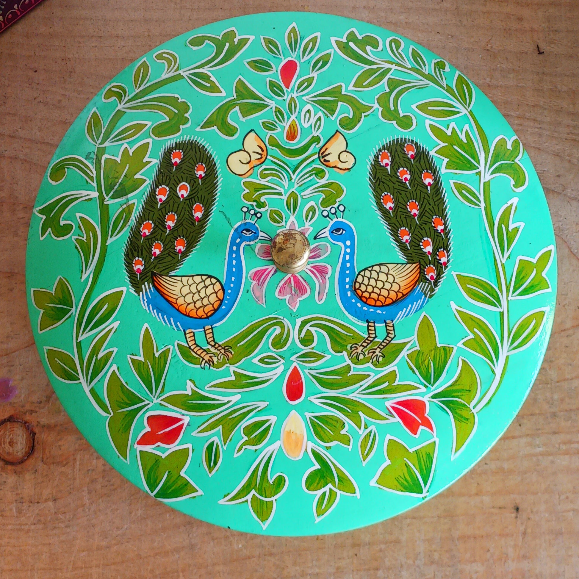 This beautiful large turquoise round Indian wooden box has been hand painted in a peacock design on the pull-off lid. It has a small round gold coloured central knob. Green leaves and pink flowers complete an appealing design.