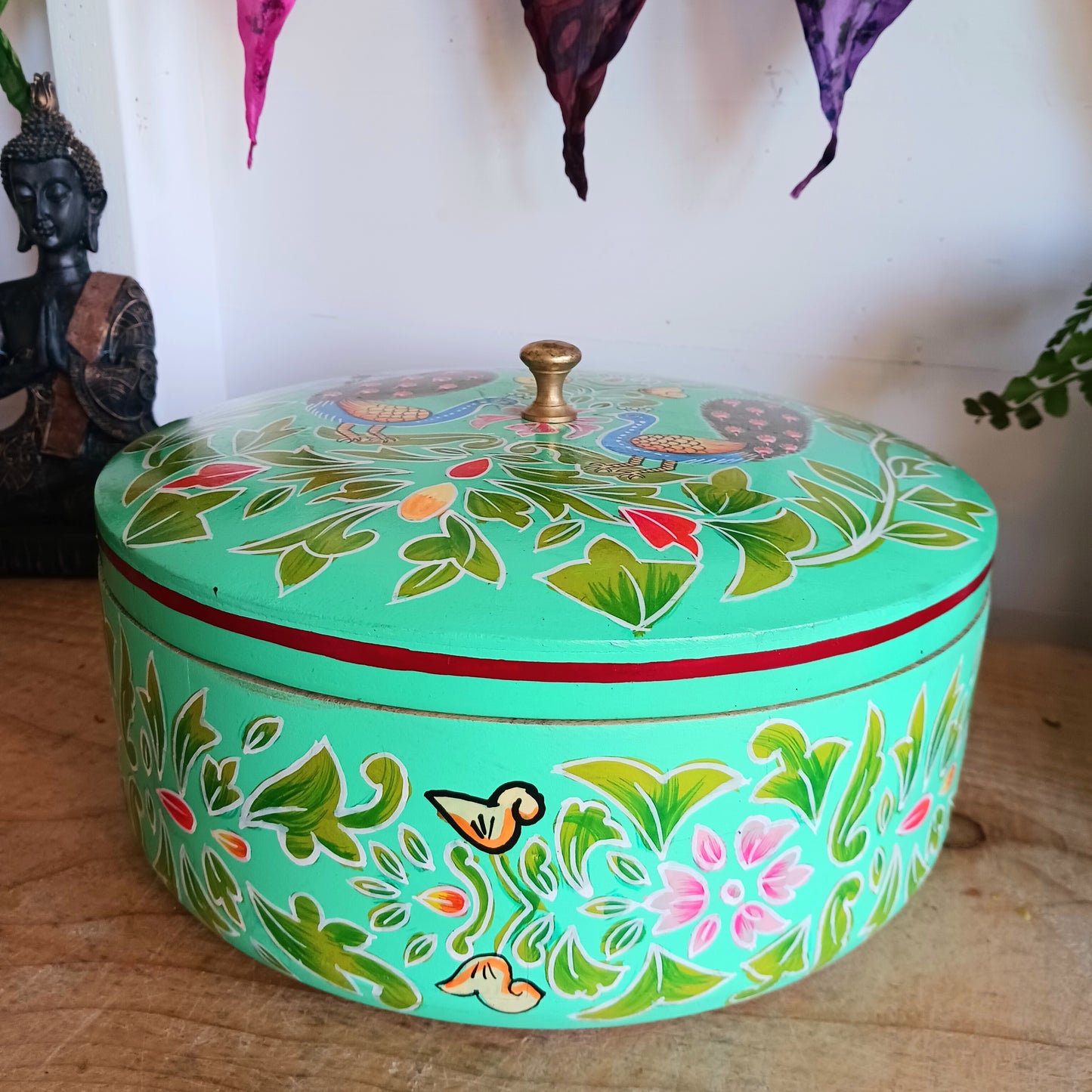 This beautiful large turquoise round Indian wooden box has been hand painted in a peacock design on the pull-off lid. It has a small round gold coloured central knob. Green leaves and pink flowers complete an appealing design.