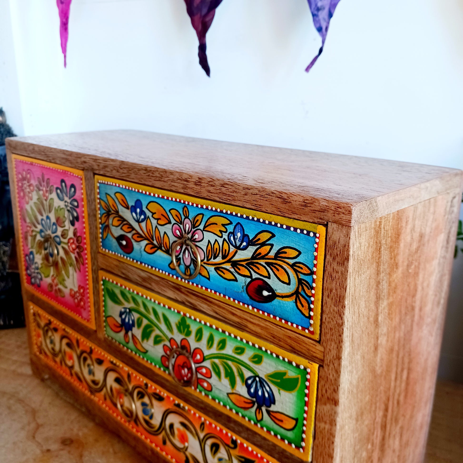 This beautiful Zaha hand painted 4 drawer mango wood chest radiates warmth and charm. Beautiful leafed vines adorn each drawer front in blue, green, red and amber with discreet central ring pull handles in gold colour. Every drawer pane is emboldened in crisp painted beading and amber.