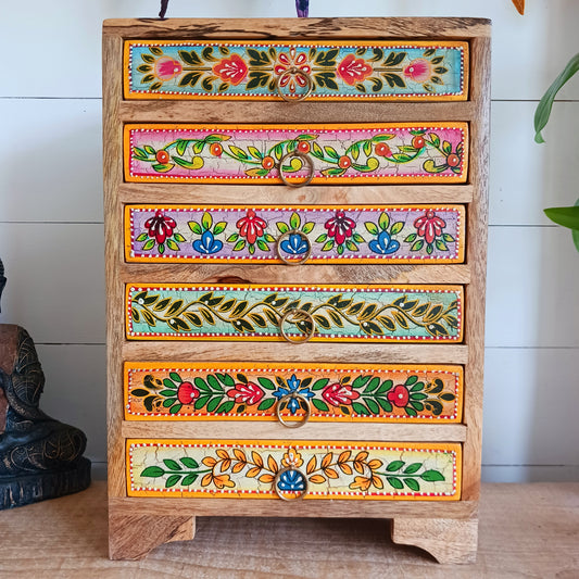 Add charm and functionality to your space with the Zaha Hand-Painted 6 Drawer Mango Wood Chest. Standing gracefully on low corner feet, this artisan-crafted piece features a vibrant blend of hand-painted leafy vines, amber-toned framing, and highlighted beading. Each drawer is uniquely designed and fitted with elegant gold-toned ring pull handles, offering a burst of colour and creativity.