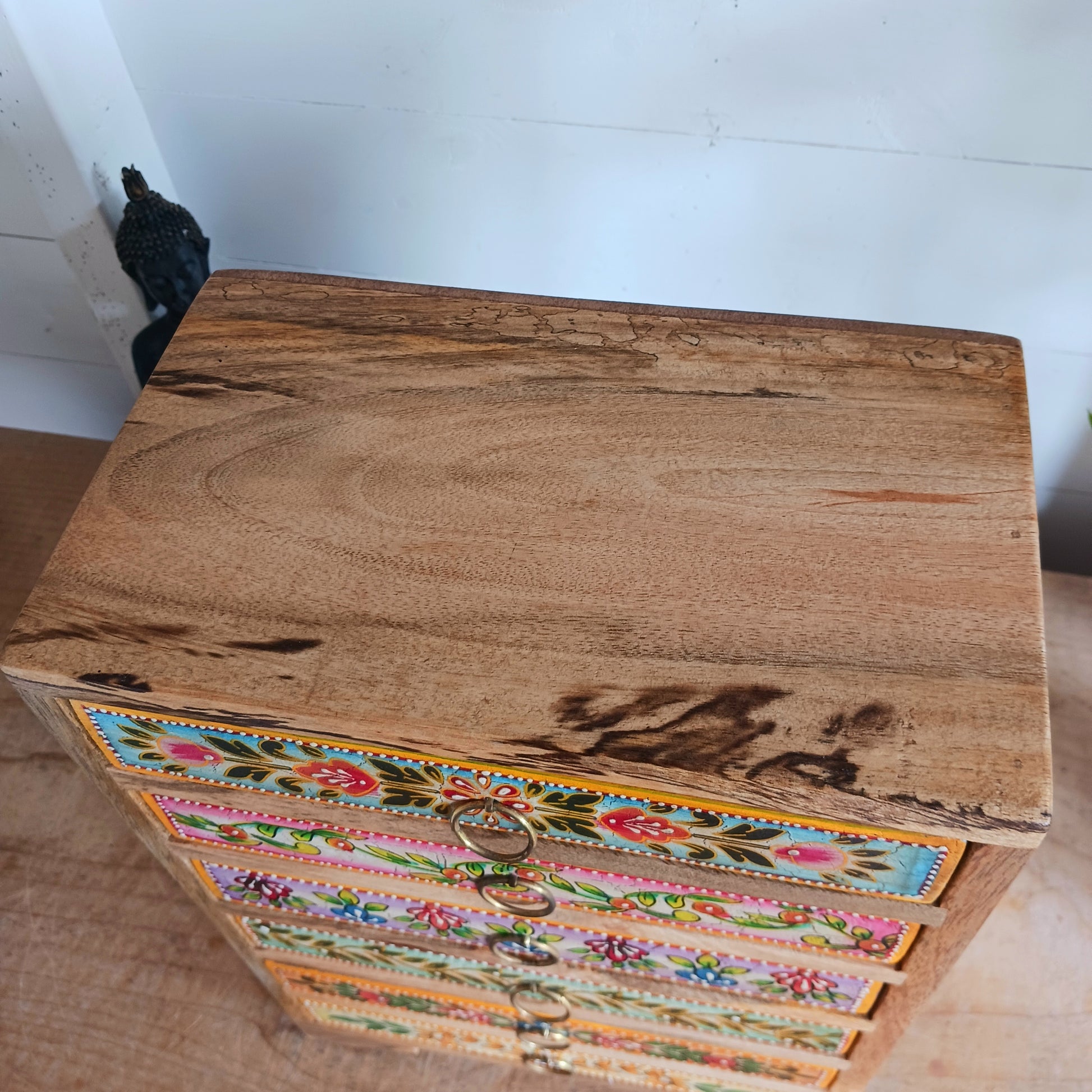 Add charm and functionality to your space with the Zaha Hand-Painted 6 Drawer Mango Wood Chest. Standing gracefully on low corner feet, this artisan-crafted piece features a vibrant blend of hand-painted leafy vines, amber-toned framing, and highlighted beading. Each drawer is uniquely designed and fitted with elegant gold-toned ring pull handles, offering a burst of colour and creativity.