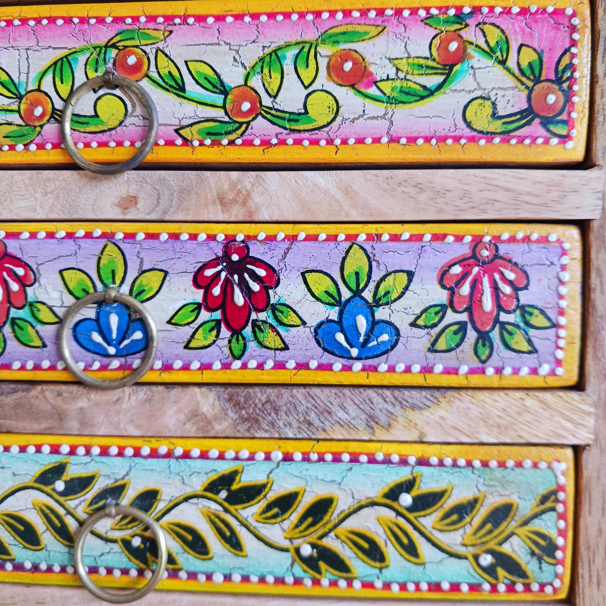 Add charm and functionality to your space with the Zaha Hand-Painted 6 Drawer Mango Wood Chest. Standing gracefully on low corner feet, this artisan-crafted piece features a vibrant blend of hand-painted leafy vines, amber-toned framing, and highlighted beading. Each drawer is uniquely designed and fitted with elegant gold-toned ring pull handles, offering a burst of colour and creativity.