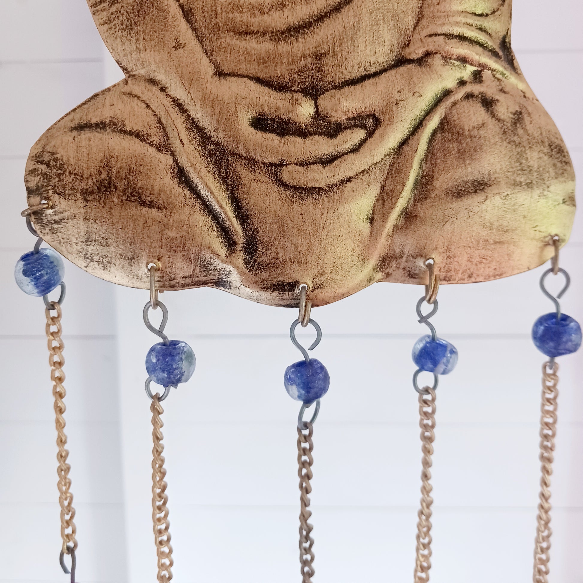 This lovely Buddha 5 bell windchime will bring serenity and cultural charm to any space.  Made in India from brass, this chime features a beautifully detailed Buddha adorned with colourful beads. 
 The five brass bells create a calming melody with every breeze, making it a perfect addition to meditation spaces, gardens, or tranquil home settings.