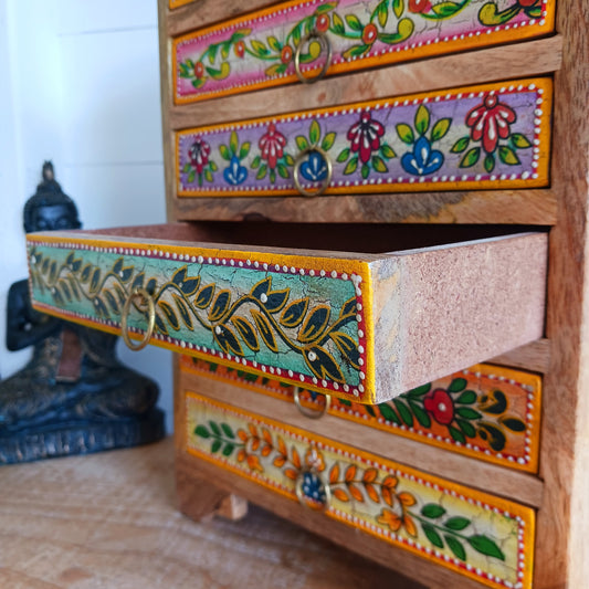 Add charm and functionality to your space with the Zaha Hand-Painted 6 Drawer Mango Wood Chest. Standing gracefully on low corner feet, this artisan-crafted piece features a vibrant blend of hand-painted leafy vines, amber-toned framing, and highlighted beading. Each drawer is uniquely designed and fitted with elegant gold-toned ring pull handles, offering a burst of colour and creativity.