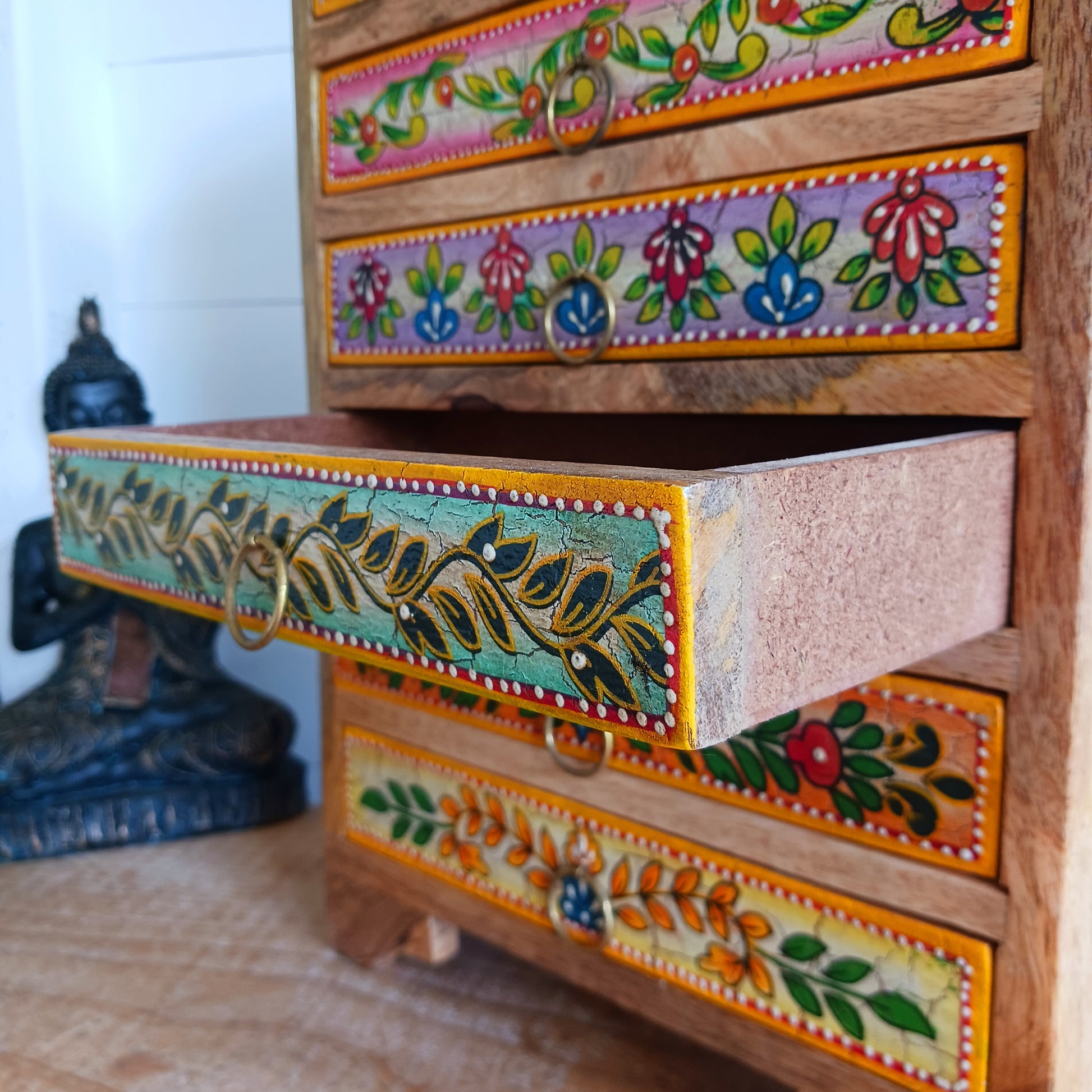 Add charm and functionality to your space with the Zaha Hand-Painted 6 Drawer Mango Wood Chest. Standing gracefully on low corner feet, this artisan-crafted piece features a vibrant blend of hand-painted leafy vines, amber-toned framing, and highlighted beading. Each drawer is uniquely designed and fitted with elegant gold-toned ring pull handles, offering a burst of colour and creativity.